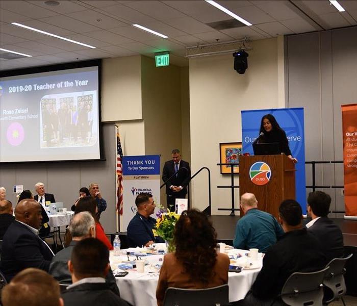 Speaker gives speech on stage while those who sit at tables watch and listen